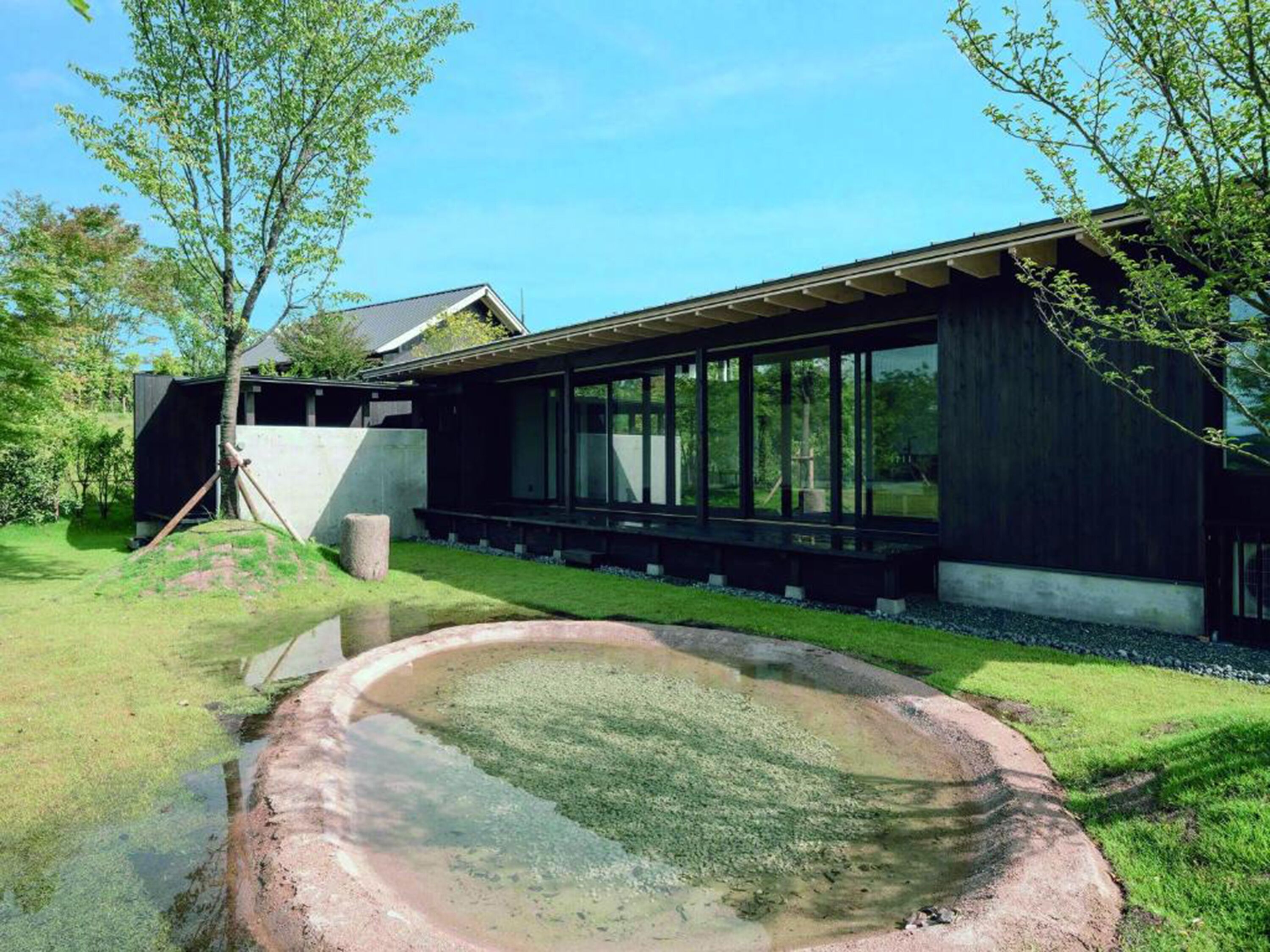 Una casa moderna con una amplia fachada de vidrio, una estructura de concreto a la izquierda y un pequeño estanque al frente.