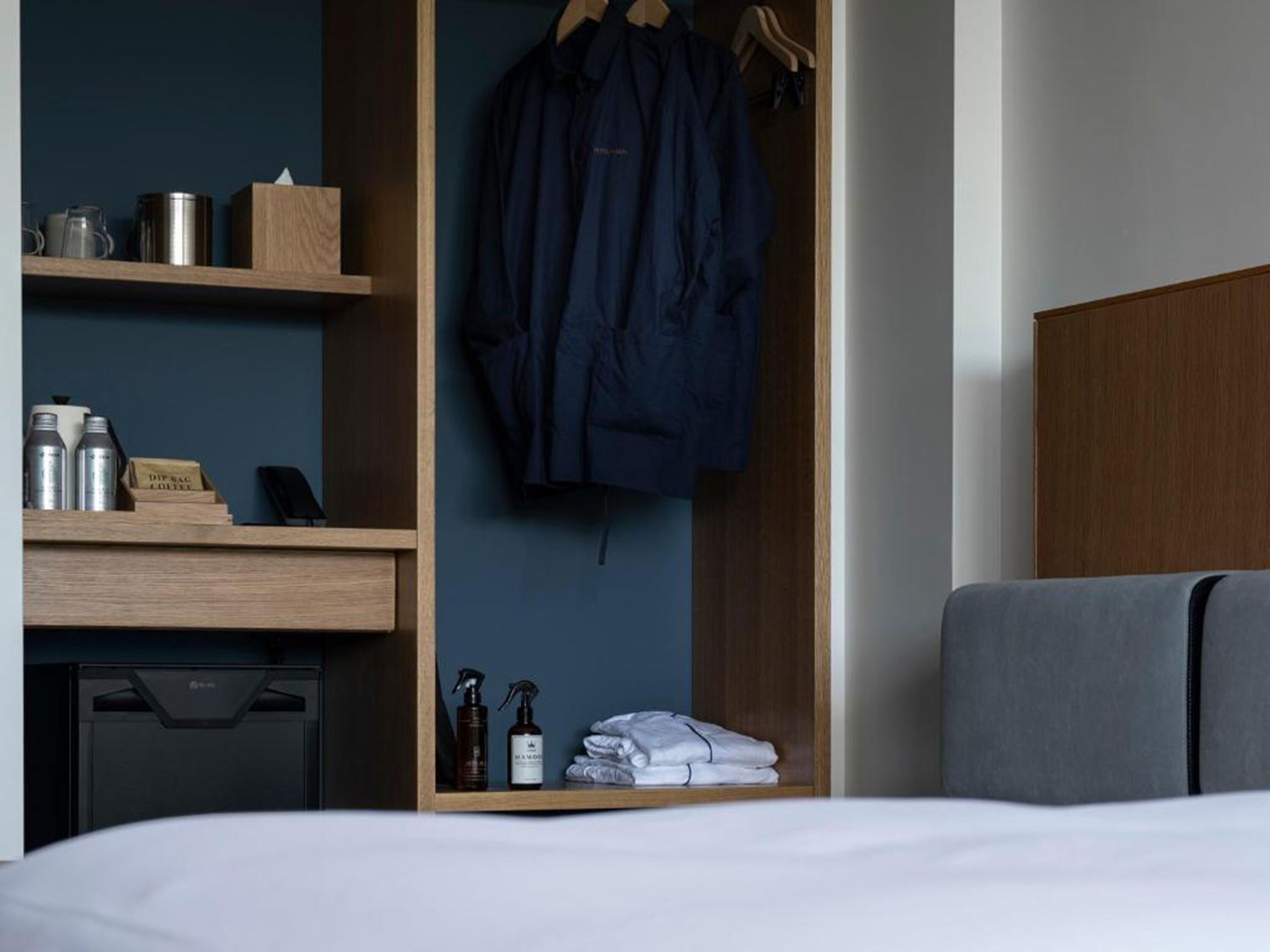 Quarto de hotel com cama, secretária de madeira com artigos de higiene, casaco pendurado e guarda-roupa.