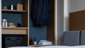 Quarto de hotel com cama, secretária de madeira com artigos de higiene, casaco pendurado e guarda-roupa.