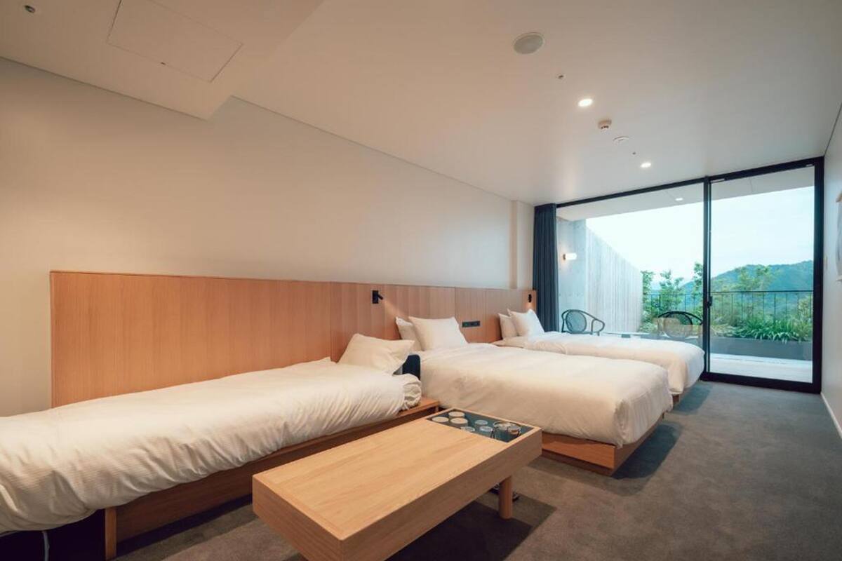 A modern hotel room with three single beds, a wooden bench, and a large window offering a view of the outdoors.