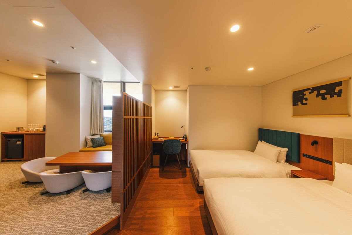 A hotel room with two beds, a desk, a chair, and a small kitchenette.