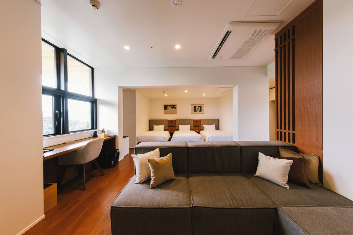 A modern hotel room with a large window, a desk, a sofa, and a bed.