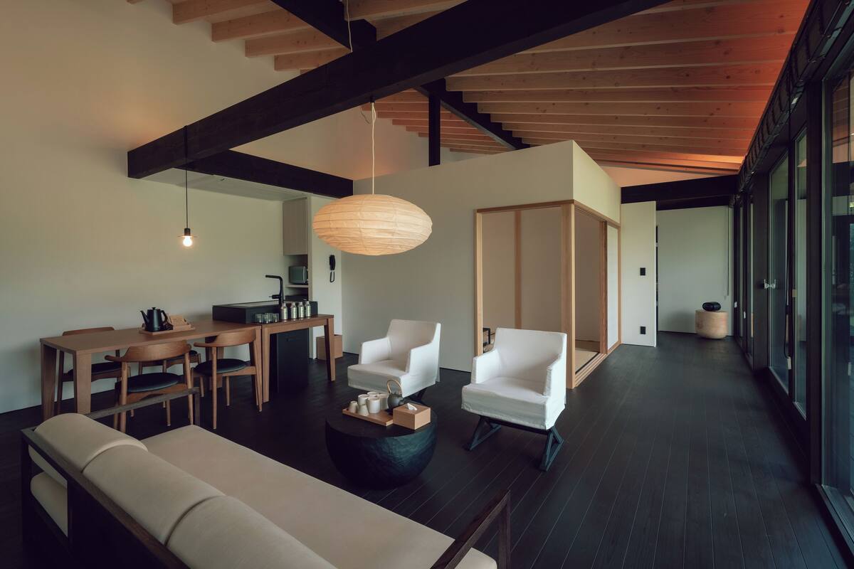 A modern living room with a sofa, two white armchairs, a wooden dining table, and a hanging light fixture.