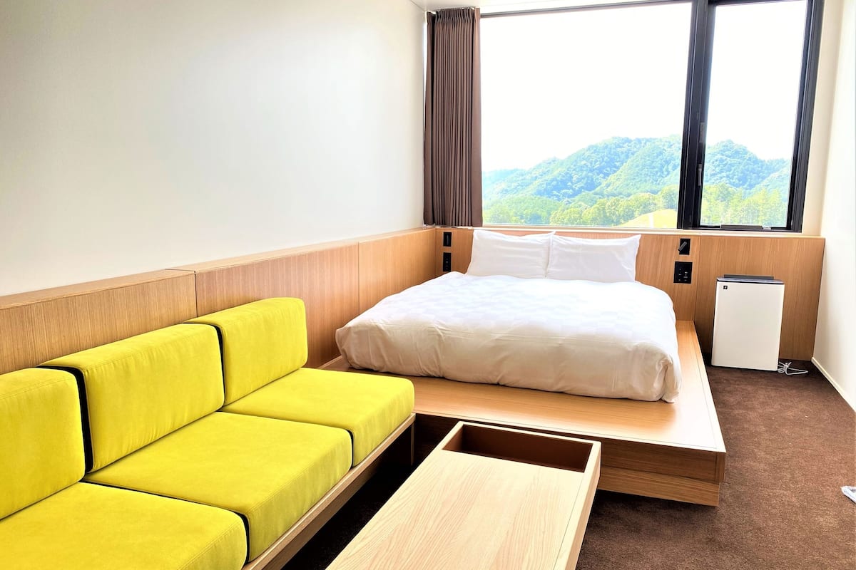 A hotel room with a bed, a yellow sofa, a small wooden table, a compact refrigerator, and a window with a view of green hills.