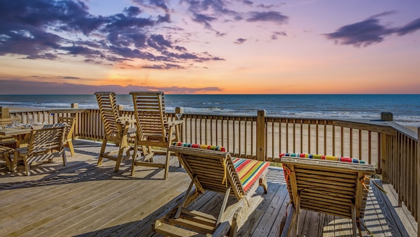 A wooden deck with lounge chairs and a table, overlooking the ocean at sunset.