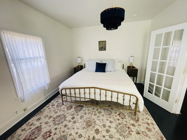 A bedroom with a bed, a chandelier, a door, and a window with curtains. - Eclectic cottage, steps from downtown (Carlton)