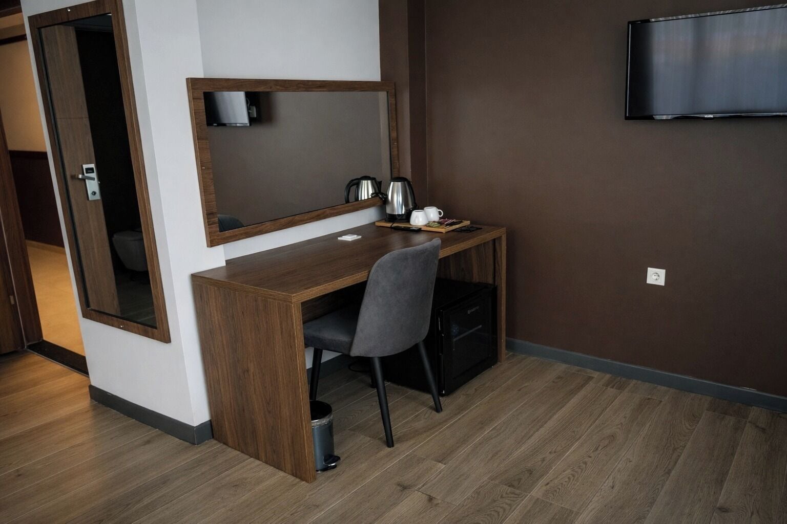 A hotel room with a wooden desk, a chair, a coffee maker, a trash can, and a television mounted on the wall.