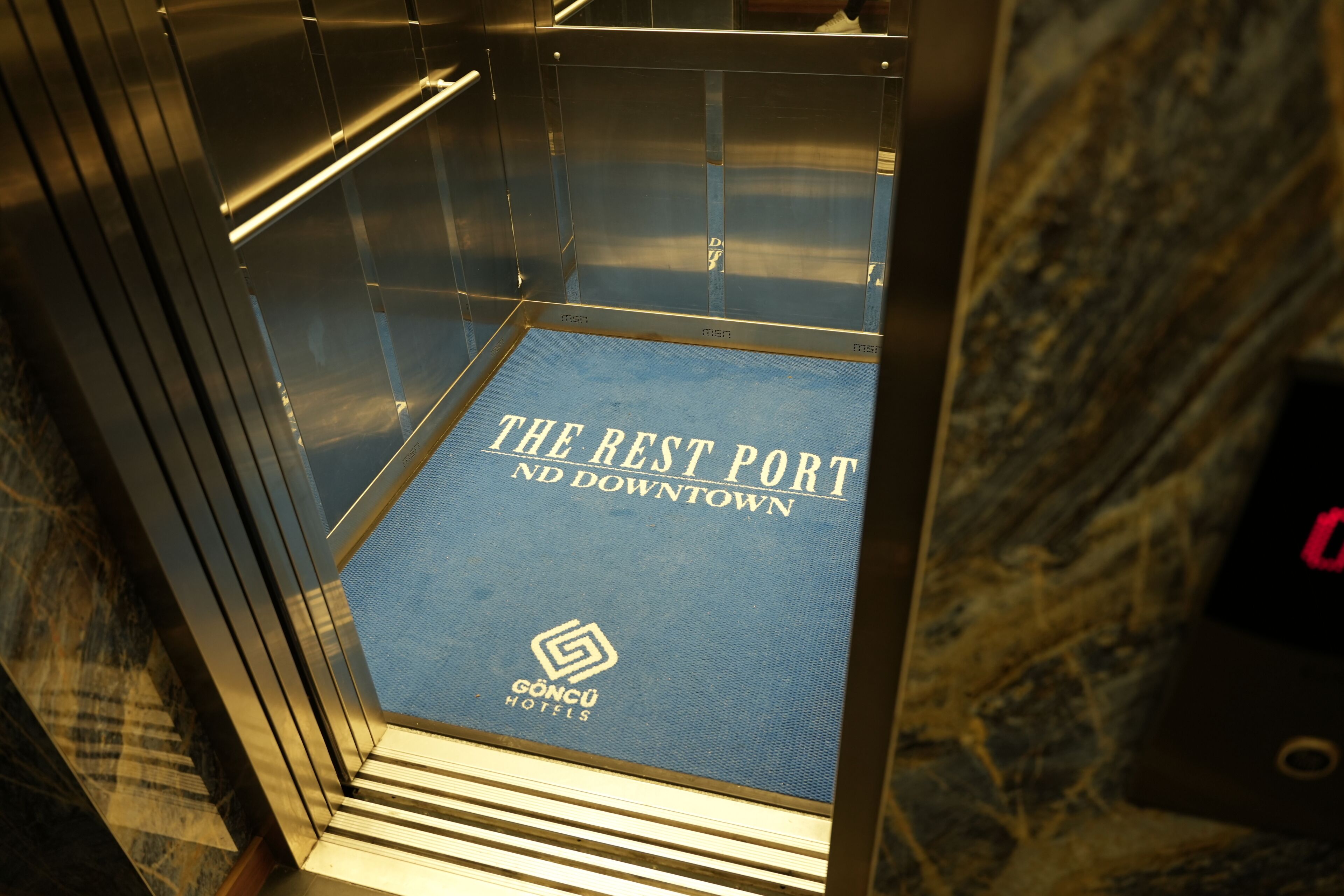 Intérieur d’un ascenseur avec un tapis bleu et l’inscription « THE REST PORT ND DOWNTOWN » ainsi que le logo « GONCU HOTELS ».