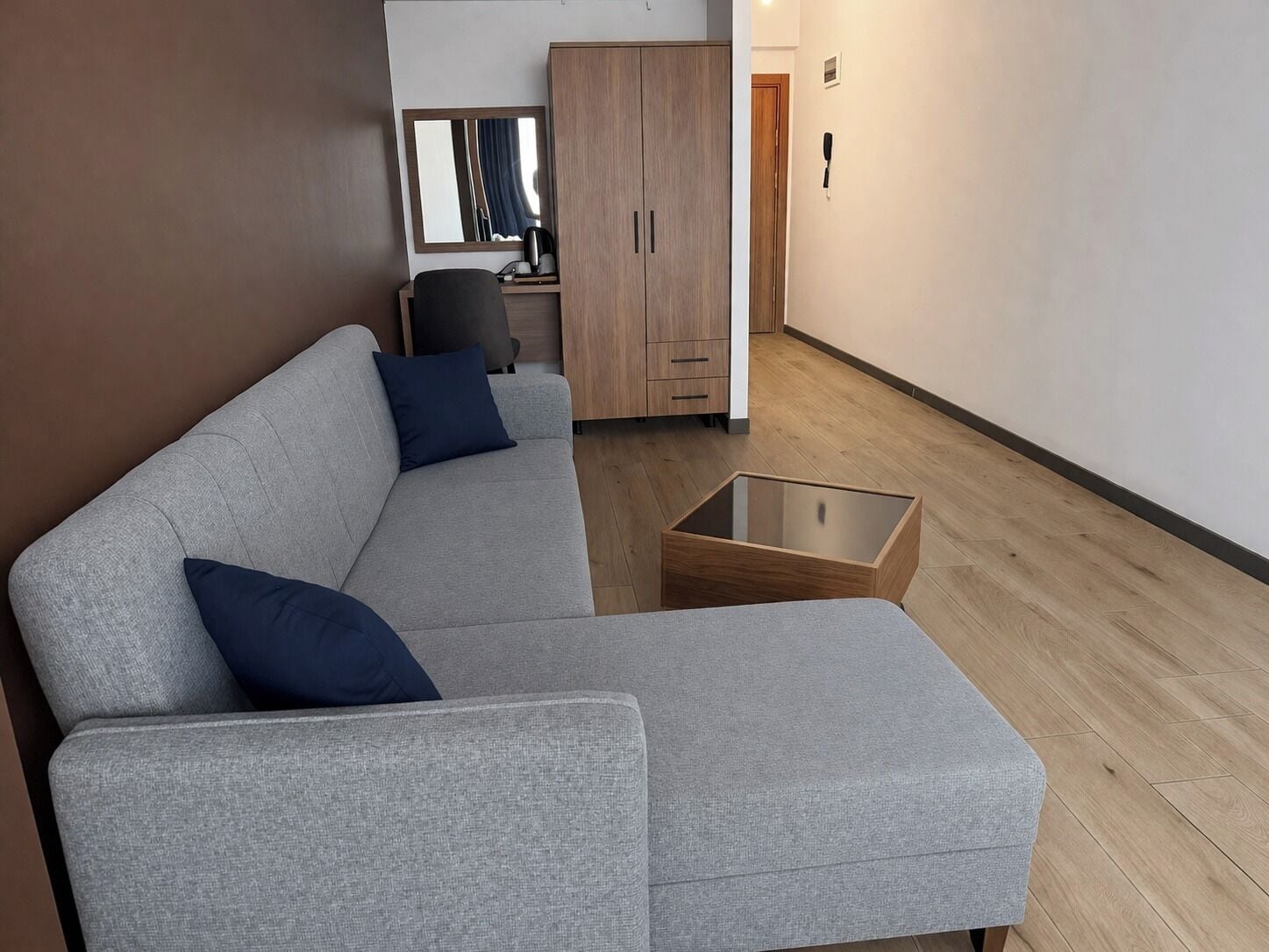 A modern living room with a grey sofa, two blue cushions, a wooden cabinet, a glass coffee table, and a wooden floor.