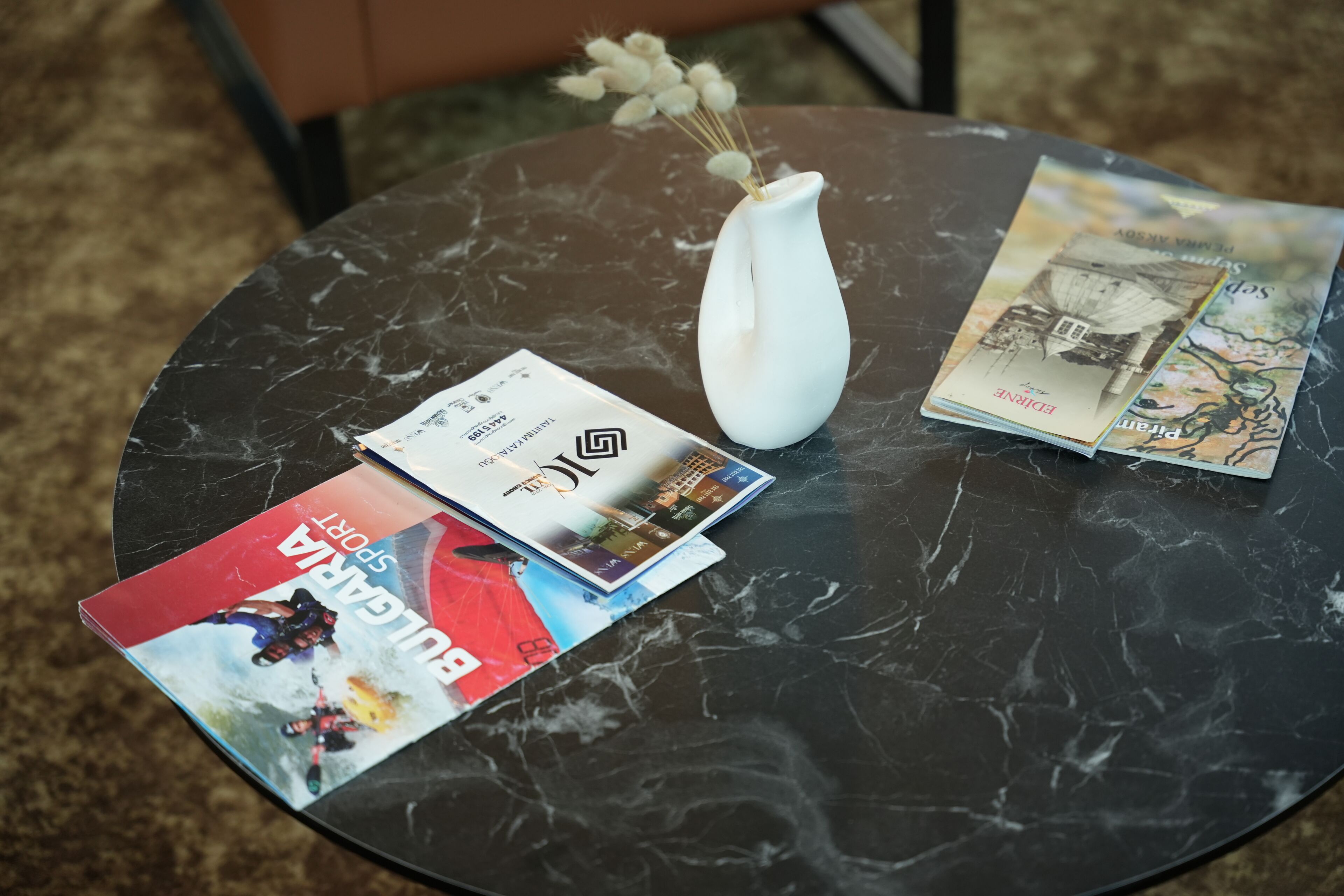 Una mesa redonda con tapa de mármol, un florero blanco con flores secas y dos revistas.