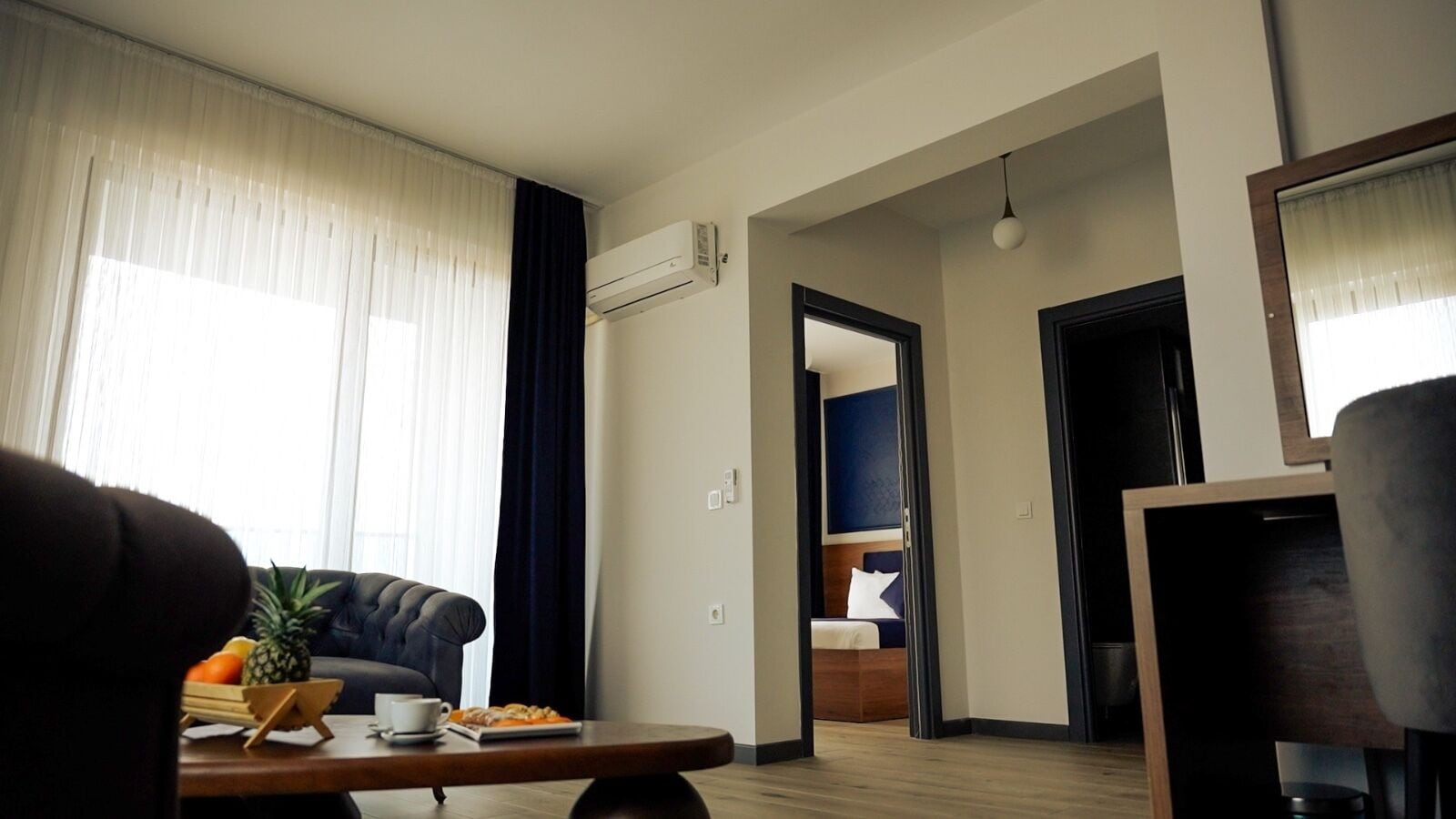 A modern living room with a sofa, a coffee table with fruits and a cup, a TV, and a hallway leading to a bedroom with a bed and a wardrobe.