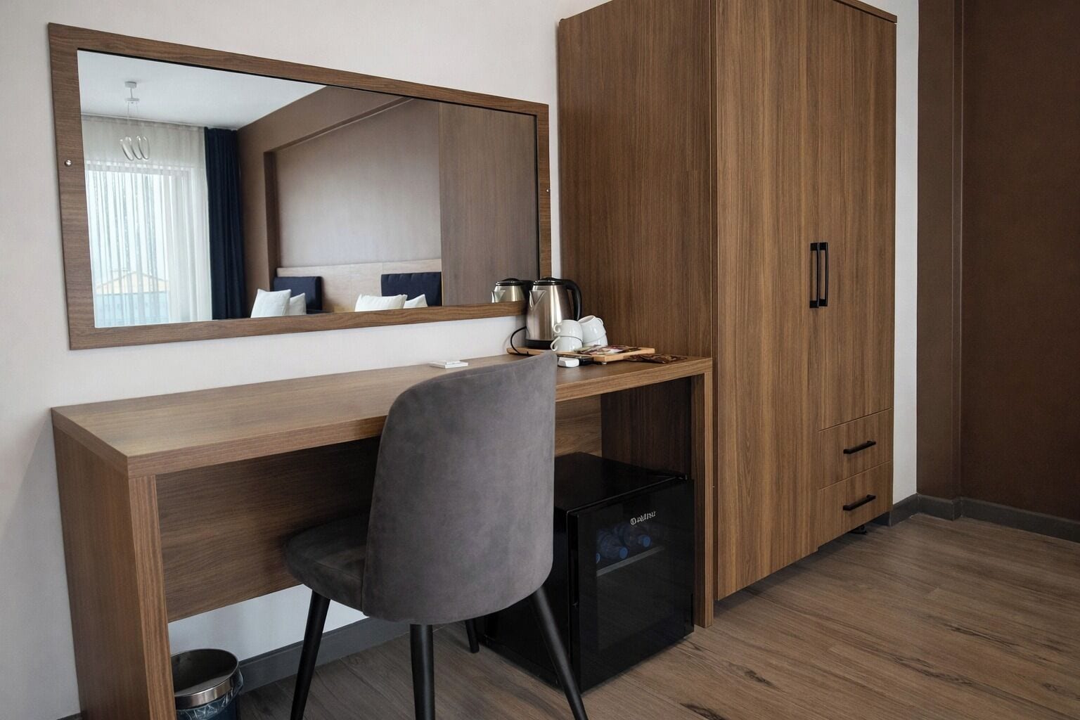 A hotel room with a wooden desk, a chair, a small refrigerator, a wardrobe, and a mirror.