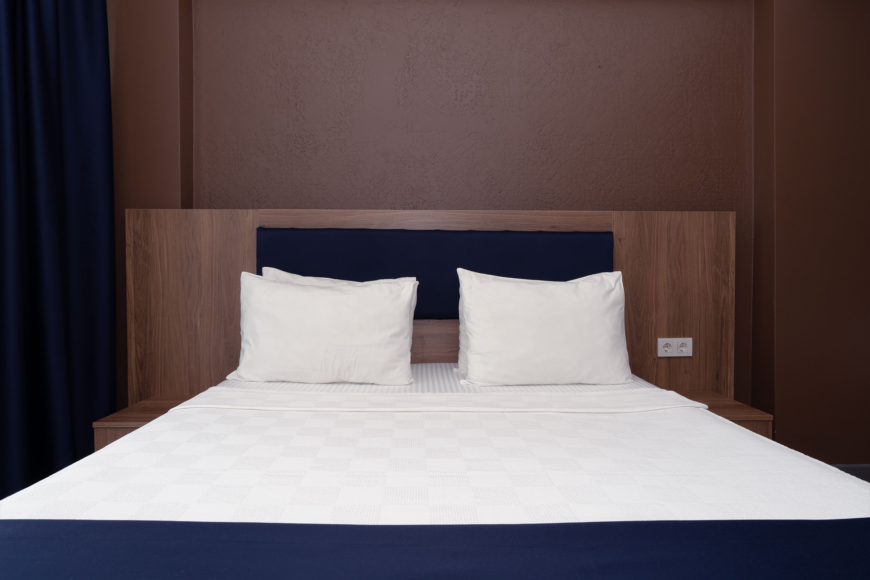 A hotel room with a bed, two pillows, a wooden headboard, and a plain wall.