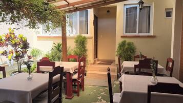 An outdoor dining area with tables and chairs, covered by a wooden pergola, and surrounded by potted plants.
