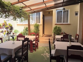 Ein Essbereich im Freien mit Tischen und Stühlen, überdacht von einer Holzpergola, umgeben von Pflanzen in Töpfen.