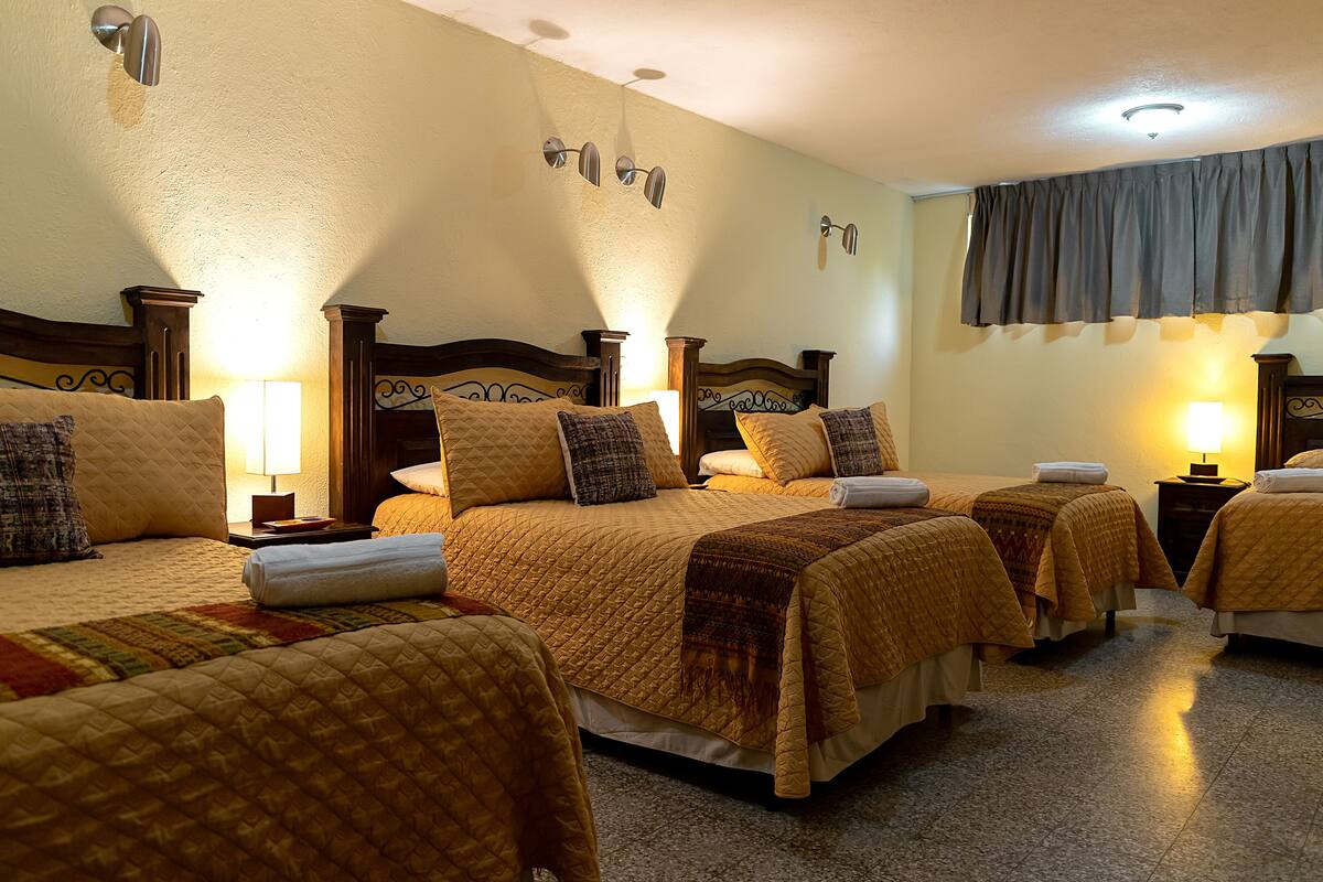 Une chambre d’hôtel avec quatre lits, chacun étant agrémenté d’une courtepointe matelassée et de coussins. On y trouve également des appliques murales et un luminaire au plafond.