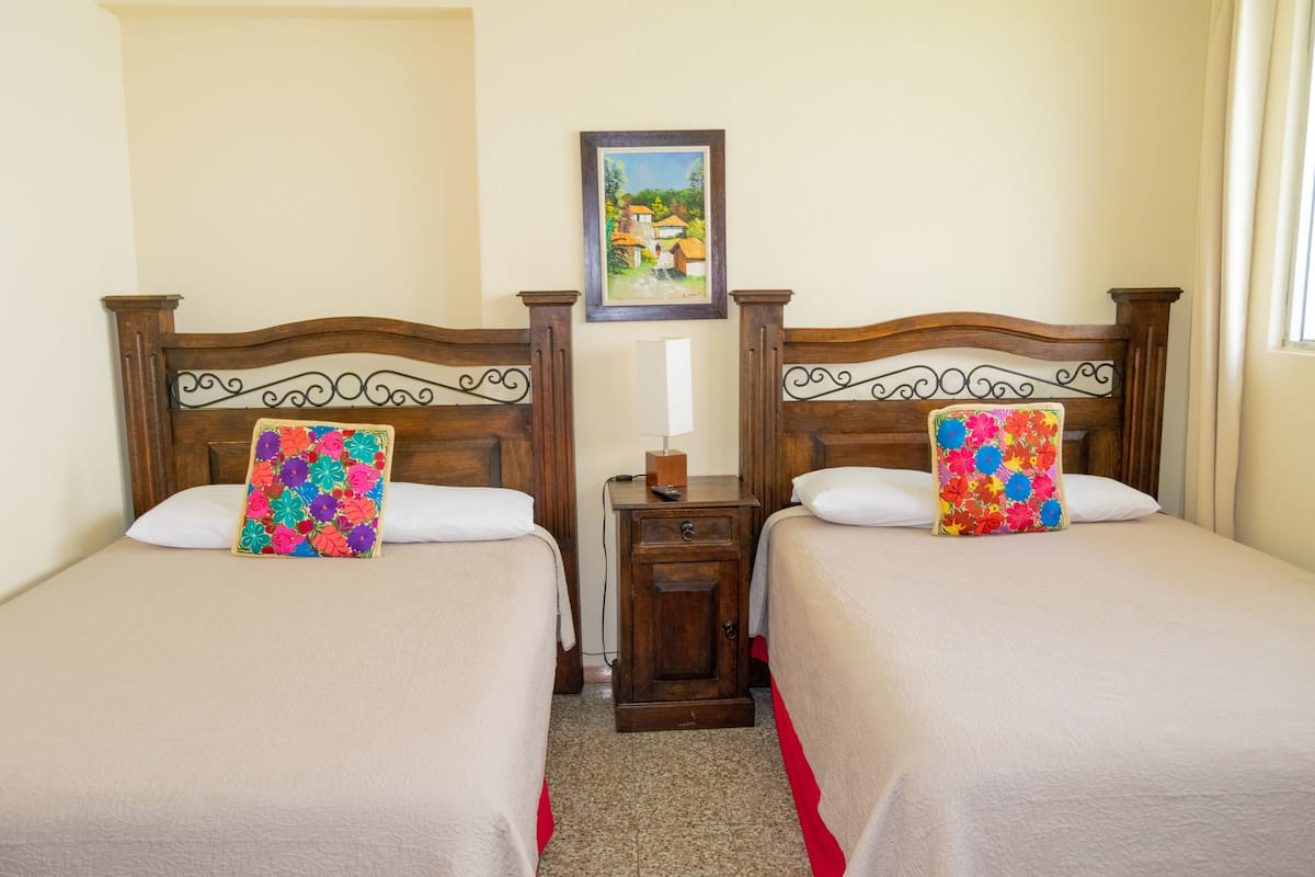 Une chambre d’hôtel avec deux lits, chacun doté d’une tête de lit en bois et d’un coussin à motifs floraux colorés. Une petite table de chevet en bois se trouve entre les deux lits, avec une lampe et une petite boîte dessus.