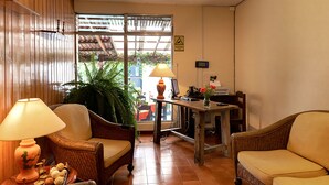 Een kamer met houten lambrisering, rieten stoelen, een tafel met een lamp en uitzicht door een raam.