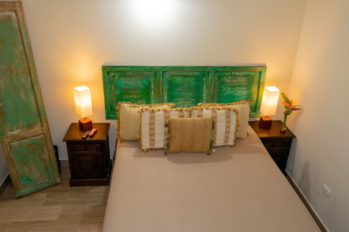 Une chambre à coucher avec une tête de lit verte, plusieurs oreillers, des tables de chevet avec des lampes et une petite table de nuit avec une télécommande.