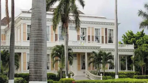 A large, two-story building with a symmetrical facade, a central entrance with a staircase, and a well-maintained garden with palm trees and hedges.