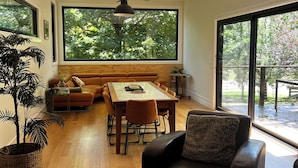 Ein geräumiger Wohnbereich mit Holzboden, einem großen Fenster mit Blick auf Bäume, einem Esstisch mit Stühlen, einer Couch und einem schwarzen Ledersessel.
