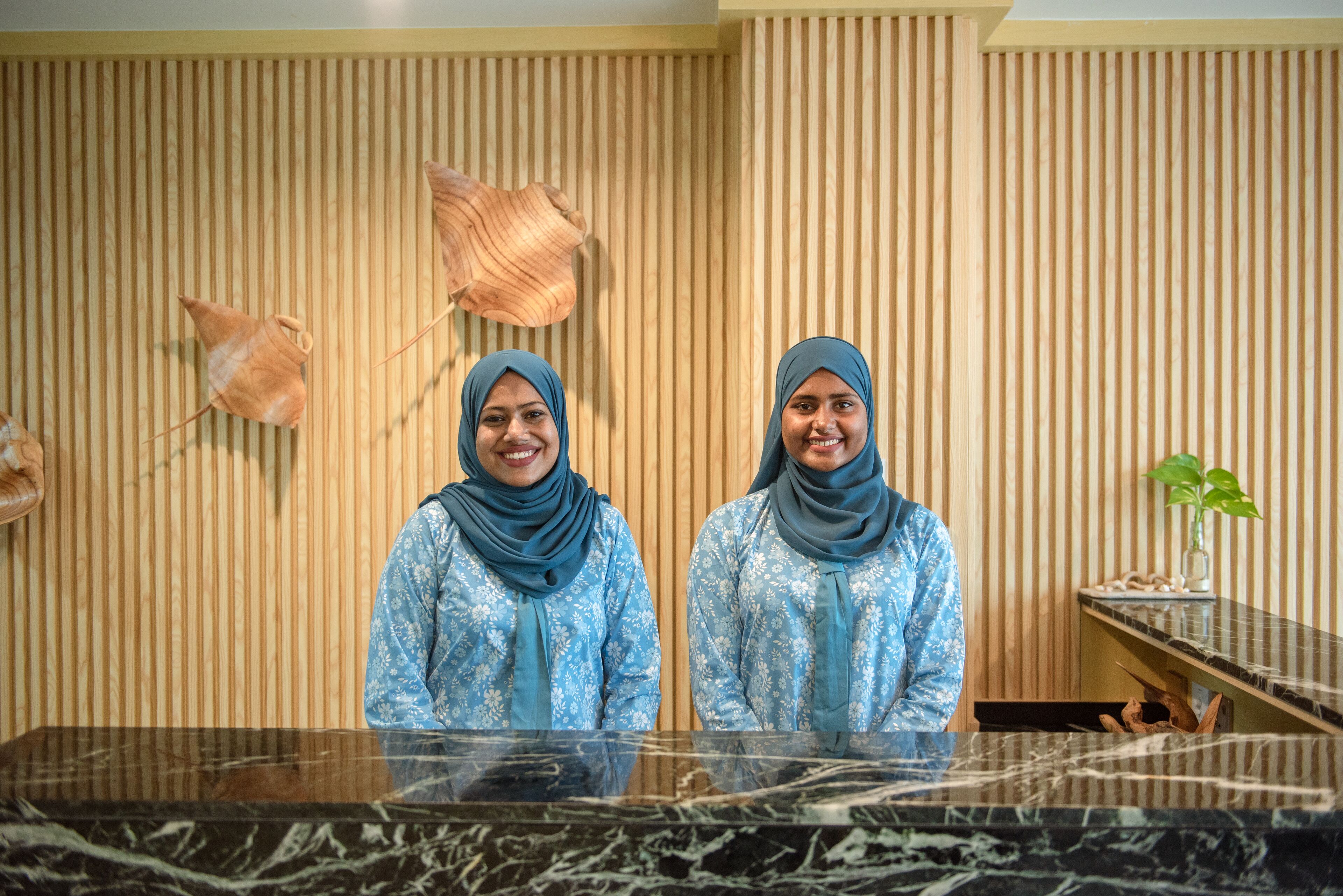 Two receptionists standing behind a marble reception desk in a room with wooden paneling and decorative pottery.