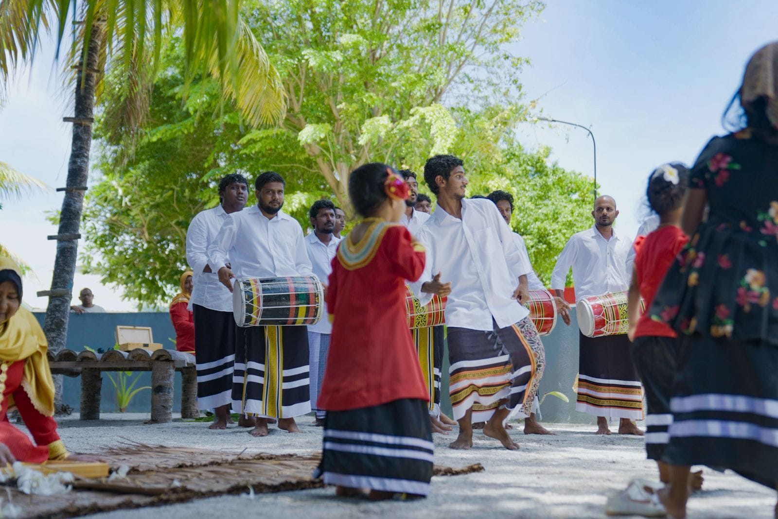 A group of people in traditional attire, some playing drums, in an outdoor setting with trees and a clear sky.