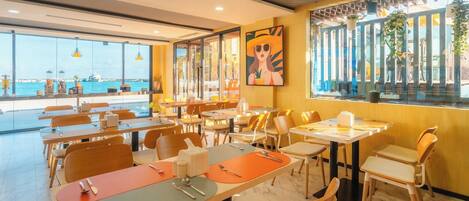 A restaurant with tables and chairs, a large window with a view of the sea, and a painting on the wall.