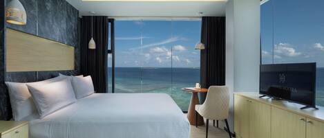 A modern hotel room with a large bed, a TV, a desk, and a view of the ocean.