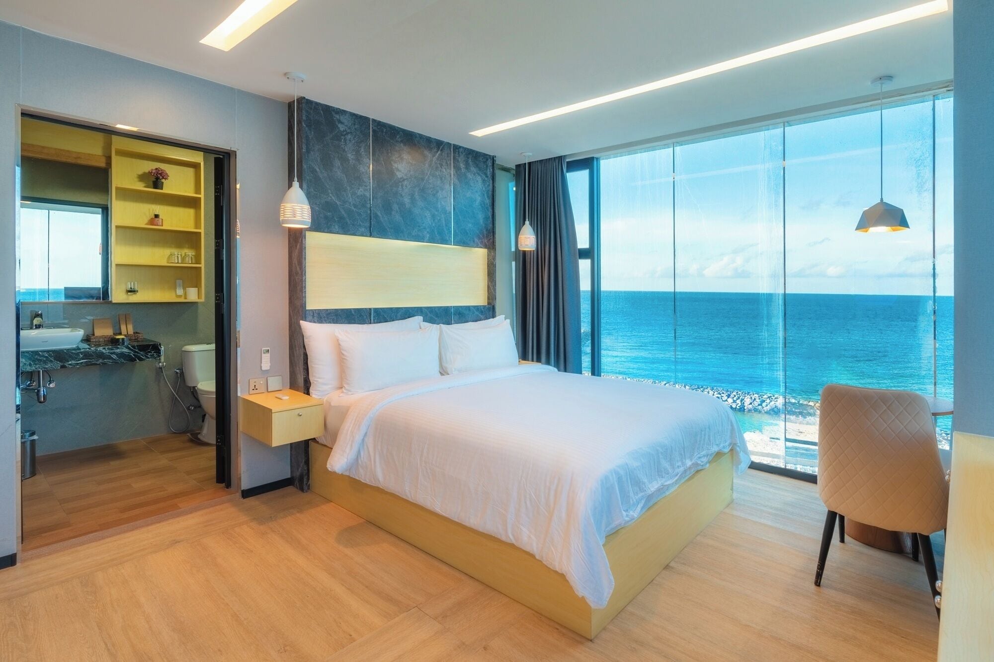 A modern hotel room with a large bed, a bedside table, a nightstand, a chair, a large window with a view of the sea, and a bathroom with a sink and mirror.