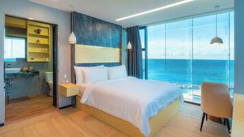 A modern hotel room with a large bed, a bedside table, a nightstand, a chair, a large window with a view of the sea, and a bathroom with a sink and mirror.