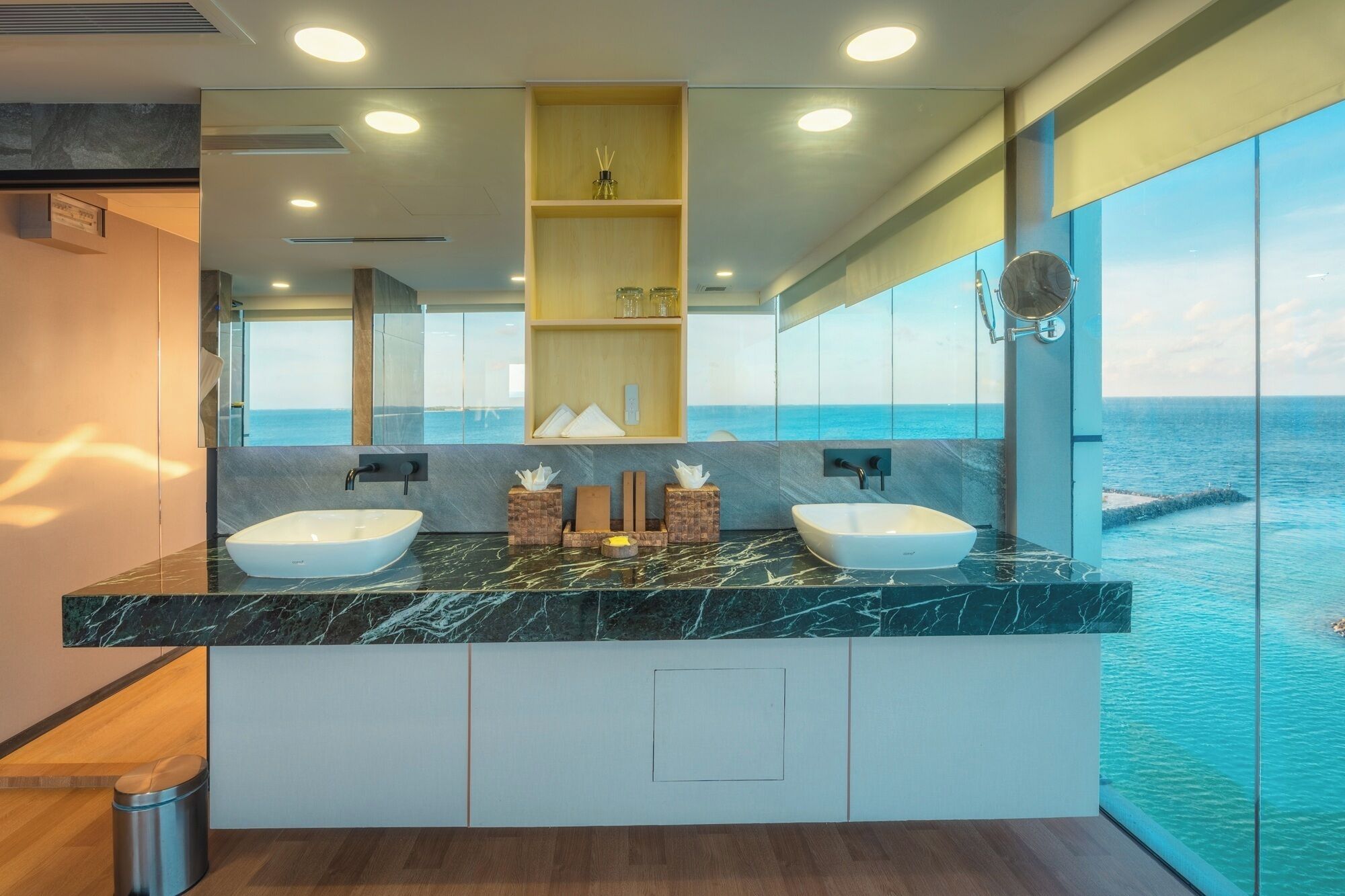 A modern bathroom with two sinks, marble countertops, and a large mirror reflecting a view of the ocean.