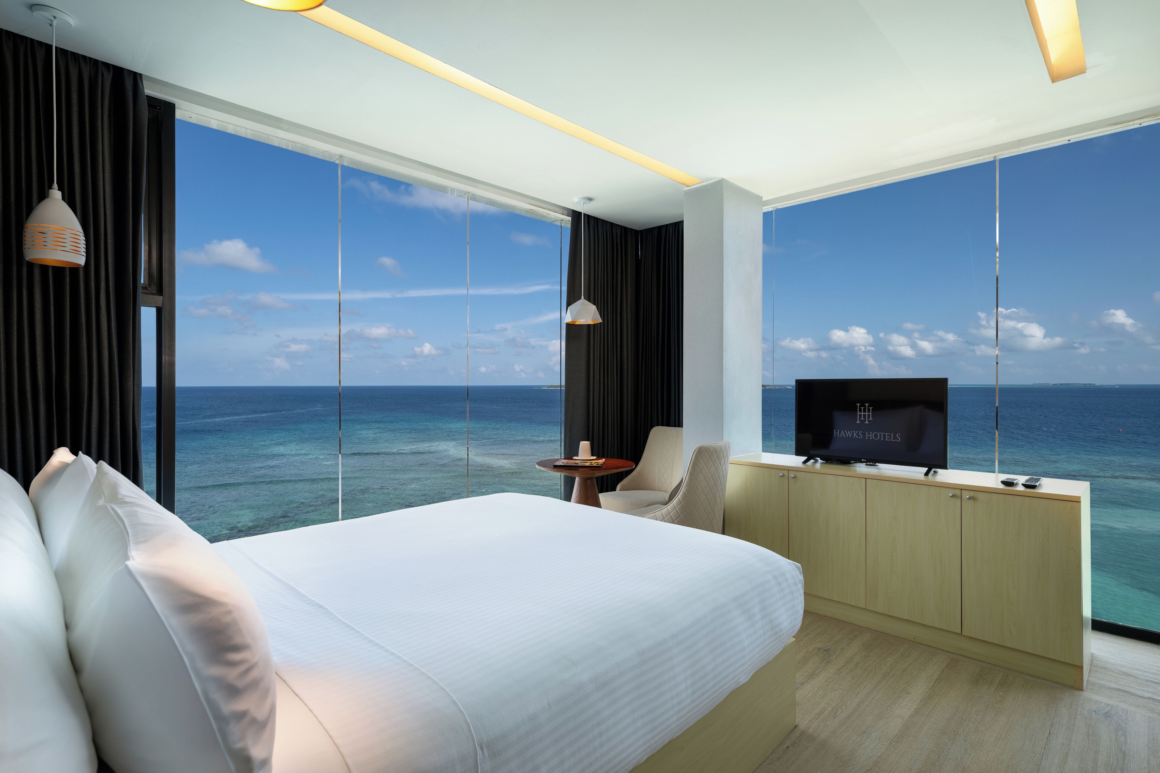 A modern hotel room with a large bed, a TV, a small table, and a view of the ocean.