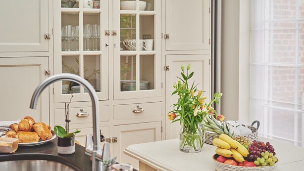 Une cuisine équipée d’un évier, d’un plan de travail sur lequel se trouve un saladier rempli de fruits, et d’un meuble avec des portes en verre contenant de la vaisselle.