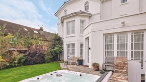 Un jacuzzi dans un jardin, avec un chemin en briques menant à une maison blanche dotée de nombreuses fenêtres.