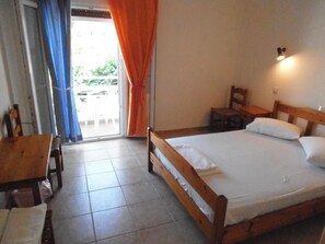 A bedroom with a wooden bed, a small table, and a chair. There is a window with curtains, and a view of the outside. - Julieta's House (Alonissos)