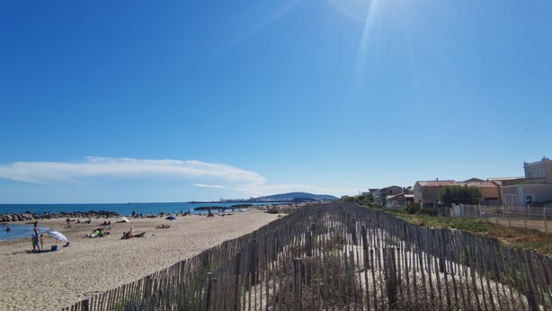 Une plage avec une clôture en bois, des personnes se promenant sur la plage et des maisons en arrière-plan.