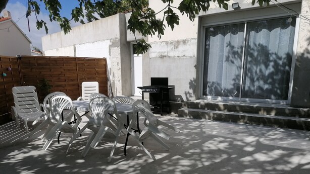 An outdoor patio with a table and chairs, a barbecue grill, and a large window. - Maison Bord de mer (Brem-sur-Mer)