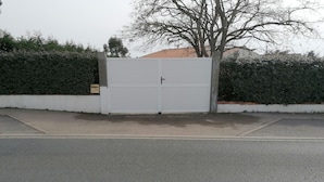 A gated entrance with a closed white gate, a concrete pathway leading to the gate, and a hedge on one side. - Maison Bord de mer (Brem-sur-Mer)