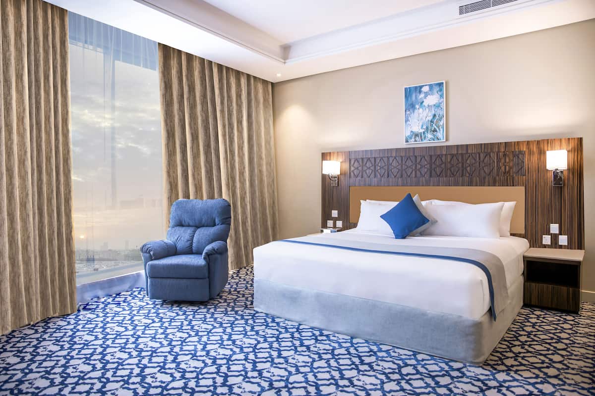 A hotel room with a large bed, a chair, a view through the window, and a patterned carpet.
