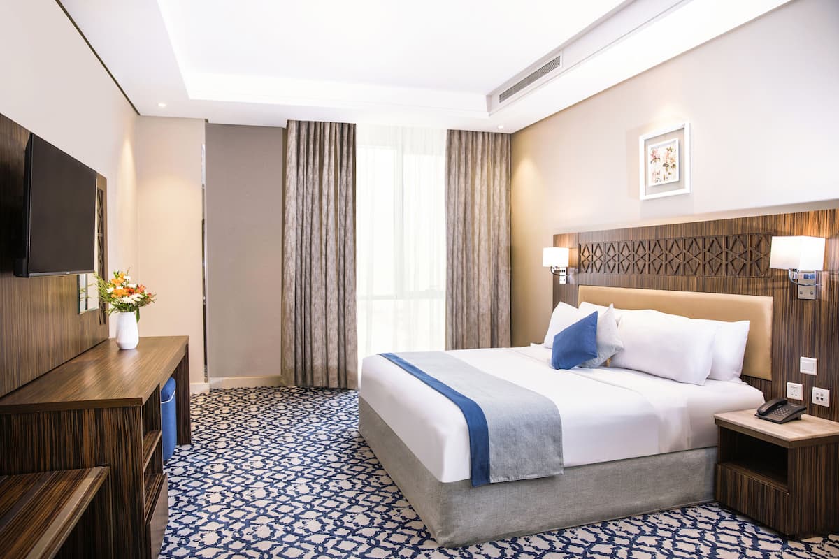 A hotel room with a large bed, a TV, a desk with a vase of flowers, and a patterned floor.