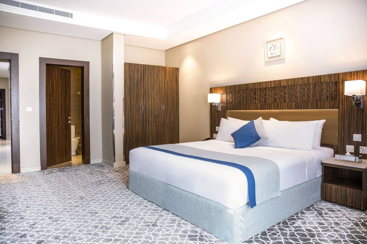 A hotel room with a large bed, a wooden wardrobe, a nightstand with a lamp, and a patterned floor.