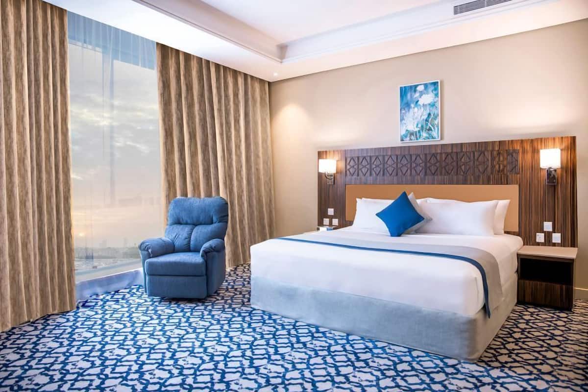 A hotel room with a large bed, a blue armchair, a patterned carpet, and a view of the city through the window.
