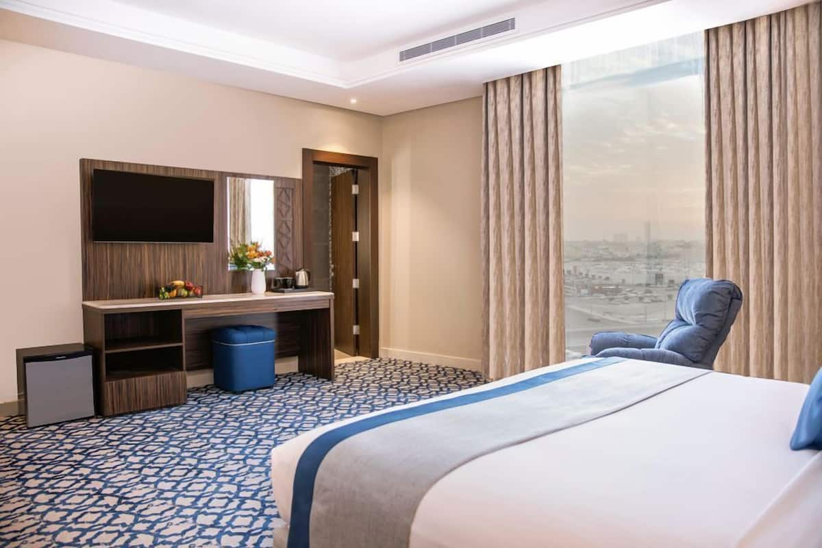 A hotel room with a bed, a desk with a flat-screen TV, a chair, and a large window with a city view.