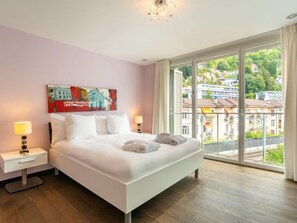 Quarto de hotel moderno com uma cama grande, duas mesas de cabeceira, um criado-mudo, um quadro na parede e uma varanda com vista para edifícios.
