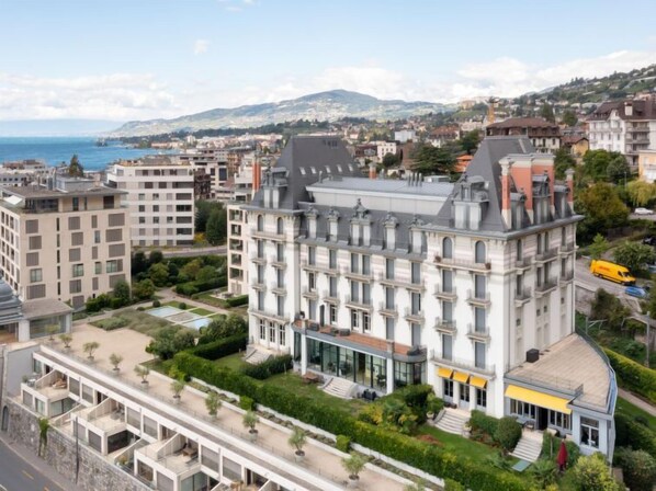A multi-story hotel with a central courtyard, surrounded by other buildings, and a view of the sea and hills in the background.