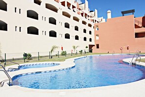 Una piscina con diseño curvo, rodeada por una valla y contigua a un edificio de varios pisos con balcones.