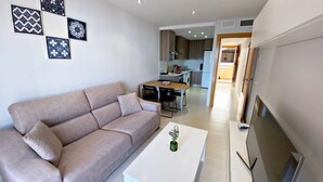 Una sala moderna con un sofá color beige, una mesita blanca y una planta verde. Se ve una cocina al fondo con armarios de madera y un microondas.