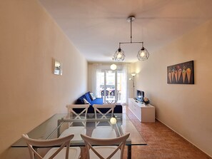 Een zithoek met een glazen tafel en stoelen, een bank, een televisie en een schilderij aan de muur.