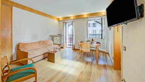 Ein Wohnzimmer mit Sofa, einem Esstisch mit Stühlen, einem an der Wand befestigten Fernseher und einem Fenster mit Blick auf Gebäude.